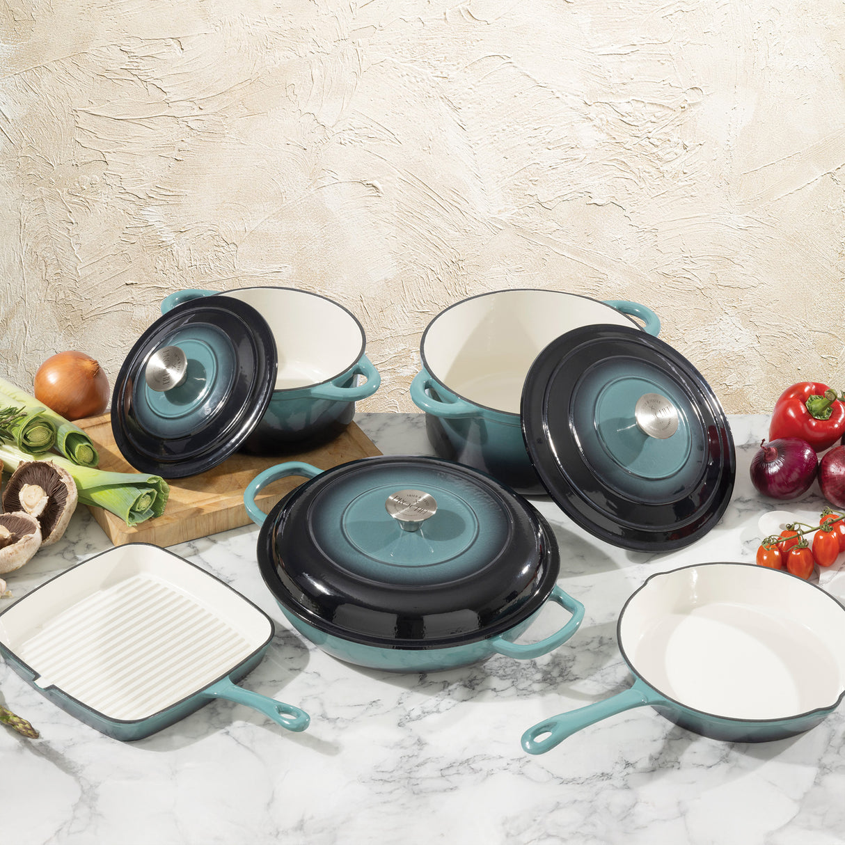 Set of teal cast iron cookware on a marble surface with vegetables.