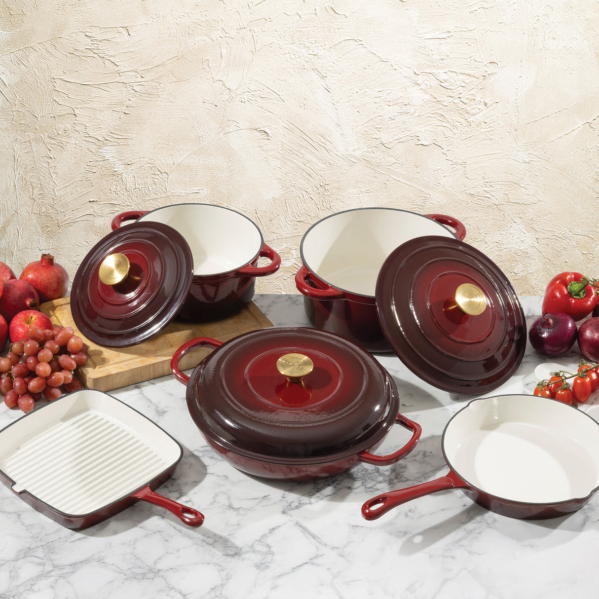 Set of deep red cast iron cookware with white interiors on a marble surface with fruits and vegetables.
