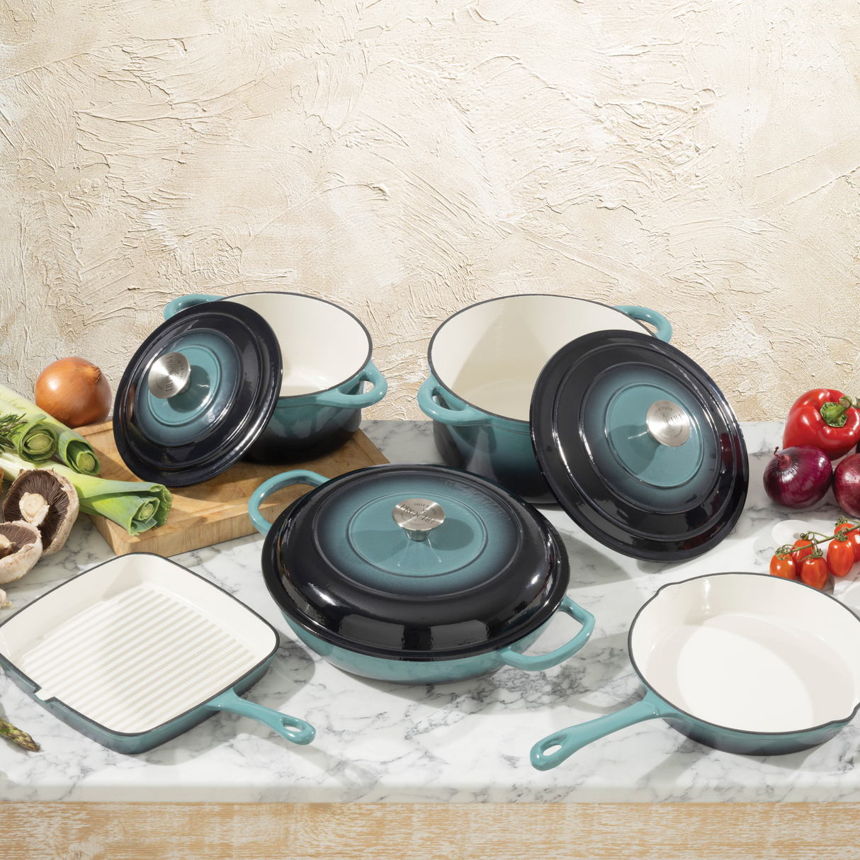 ProChef 5-piece teal cast iron cookware set with white interiors, including two round casseroles with lids, one shallow casserole with lid, a grill pan, and a skillet, displayed on a marble countertop with fresh vegetables.