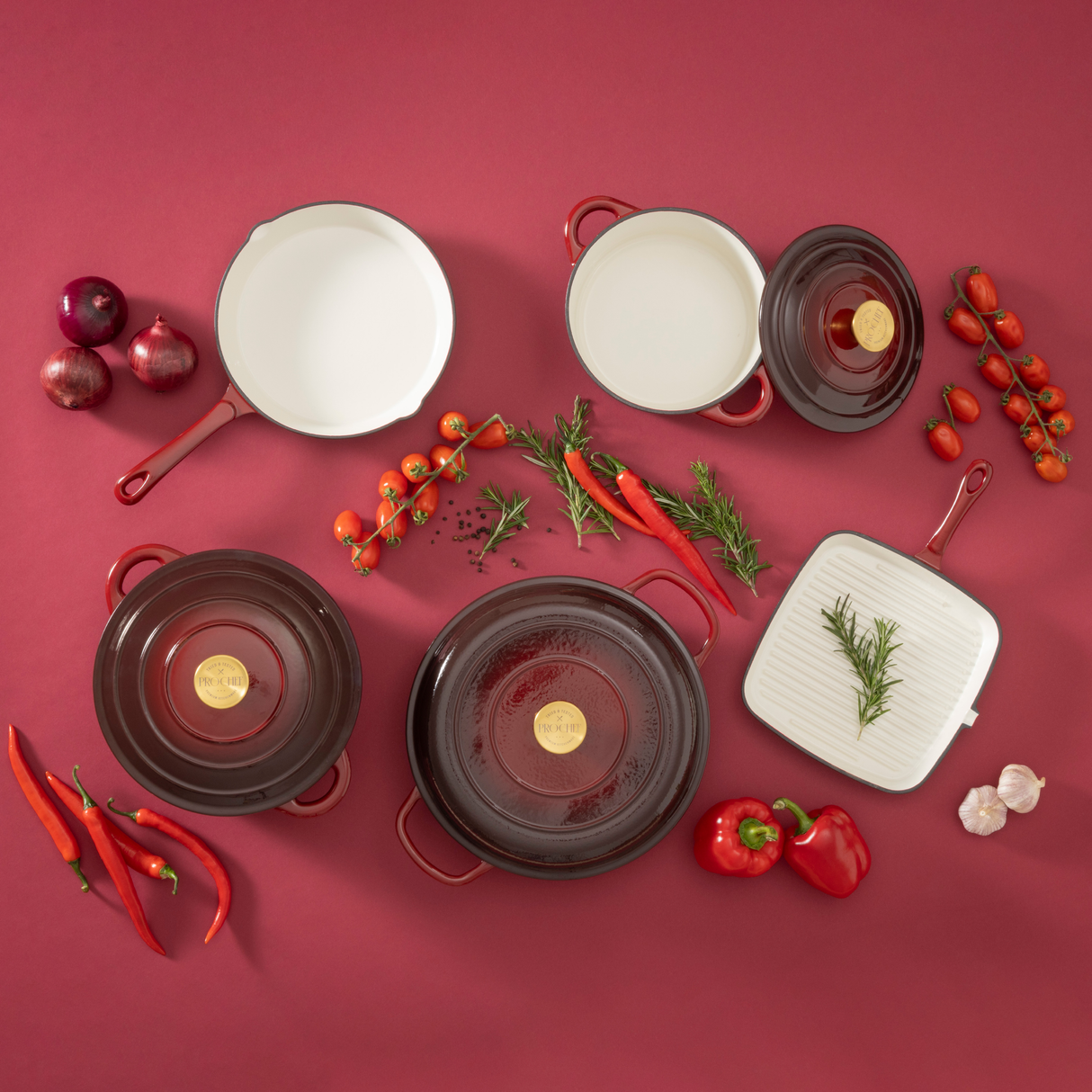 Set of ProChef deep garnet cast iron cookware on a red background with vegetables and herbs.