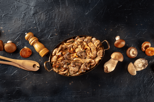 Dried mushrooms in a pan on a dark textured surface with whole mushrooms around