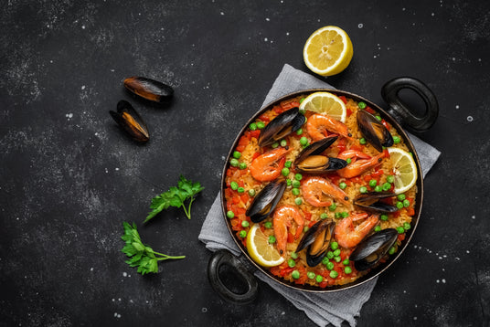 Seafood paella in a pan on a dark surface with lemon wedges and mussels.