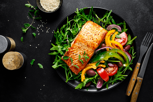 Salmon fillet on a bed of greens with vegetables on a black plate, set against a dark background.