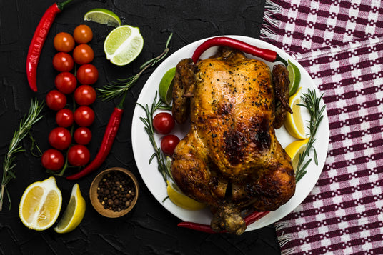 Roasted chicken on a white plate with herbs, tomatoes, and lemon slices on a dark surface.
