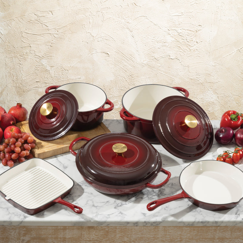 Set of red cast iron cookware with white interiors on a marble surface with fruits and vegetables.