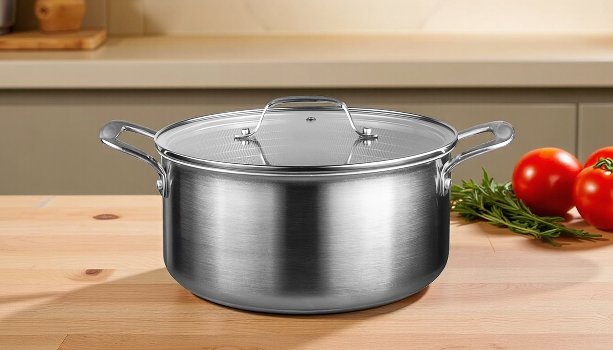 Stainless steel pot with lid on a wooden surface with tomatoes and herbs in the background
