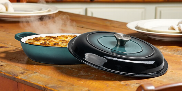 ProChef gradient teal cast iron oval casserole dish with lid beside it, filled with a baked dish and steaming on a wooden kitchen table.