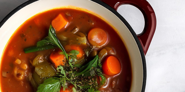 Vegetable soup with carrots and greens in a red pot on a white background