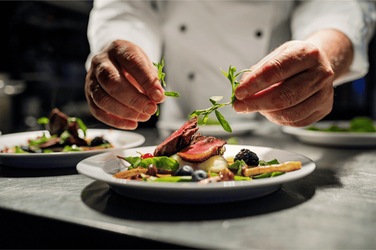 Chef in white uniform plating gourmet steak with fresh herbs and vegetables in a fine dining kitchen.