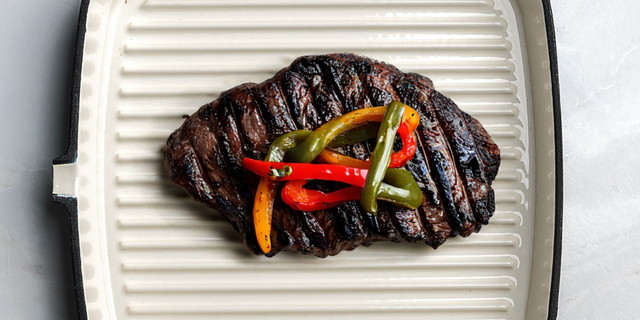 ProChef cast iron grill pan with white enamel interior, holding a grilled steak topped with roasted red, yellow, and green peppers.