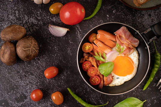 Breakfast dish with eggs, sausages, and tomatoes on a dark surface with ingredients around.
