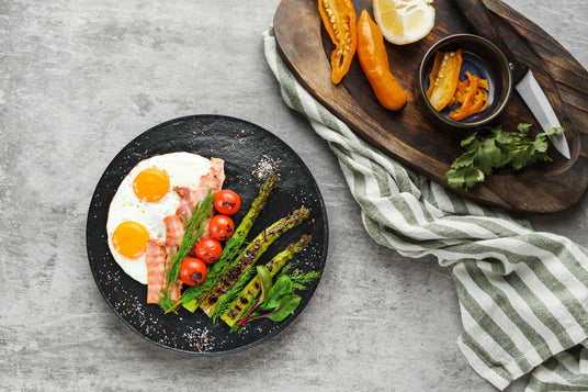 Breakfast plate with eggs, bacon, and vegetables on a gray surface with a wooden cutting board.