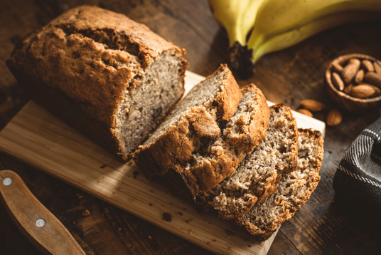Loaf of banana bread with a few slices cut, on a wooden board with bananas and nuts in the background.