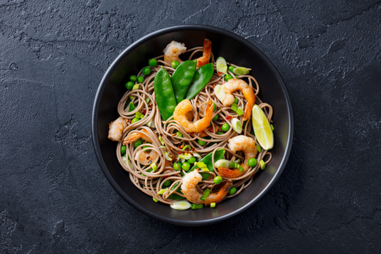 Soba noodle dish with shrimp and vegetables in a black bowl on a dark background