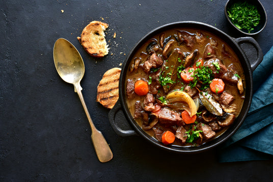 Stew with vegetables in a black pot on a dark surface with a spoon and bread slices.