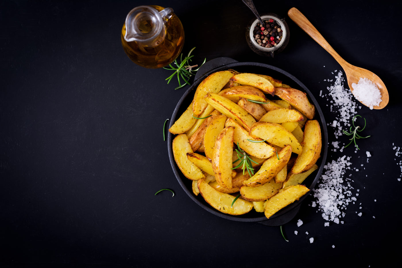 Baked potato wedges in a black bowl on a dark surface with seasonings.