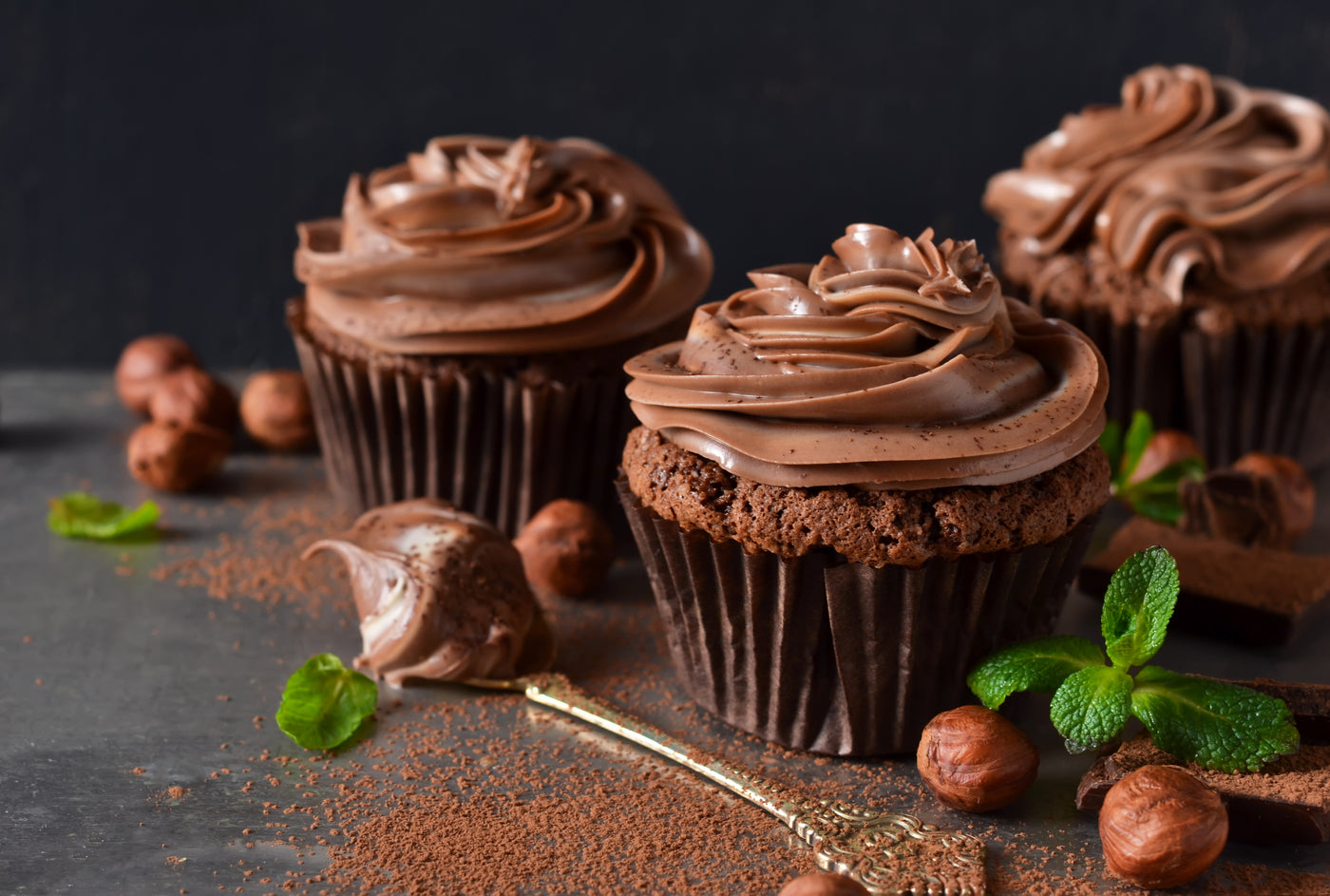 Chocolate cupcakes with frosting on a dark surface with chocolate shavings and nuts.