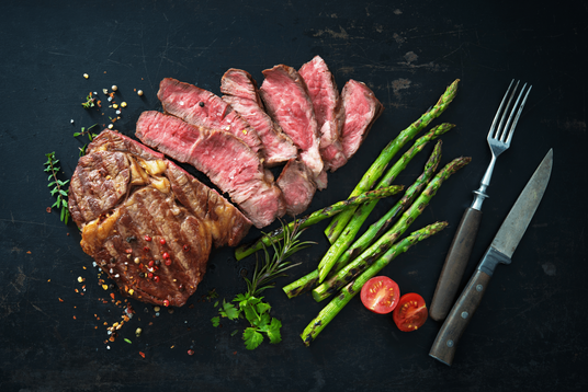 Sliced steak with asparagus, tomatoes, and herbs on a dark surface with a fork and knife.